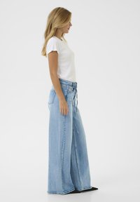 Woman with long blonde hair stands sideways wearing a white short-sleeve top and light blue wide-leg jeans with black shoes.