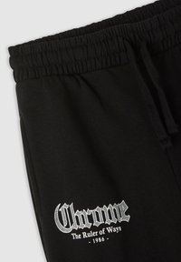 Czarne dresy z elastycznym paskiem w talii, sznurkiem do regulacji i białym logotypem "Chrome the Ruler of Ways 1986" na dolnej części lewej nogawki.