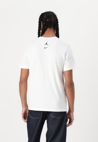 Bílý tričko s krátkým rukávem, na kterém je na horní zadní části potištěno černé logo Jordan a znak Nike, vyrobené z hladkého a lehkého materiálu.