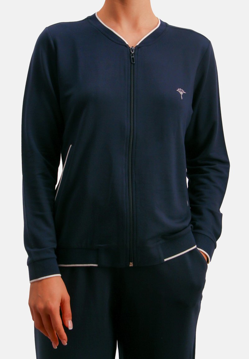 Chaqueta deportiva azul marino con cremallera, confeccionada en tejido liso, con ribete blanco en el cuello y los puños, bolsillos laterales y un bordado de logo sutil.