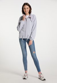 Světle šedá mikina na zip s vyšitým srdcem, stahovací pas a uvolněný střih, spárovaná s modrými slim džíny a sportovními teniskami.