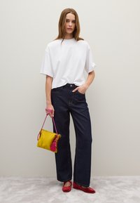 Camiseta blanca de gran tamaño, jeans de tiro alto azul oscuro, bailarinas rojas y un bolso amarillo con correa rosa y detalle de borlas. Fondo neutro.
