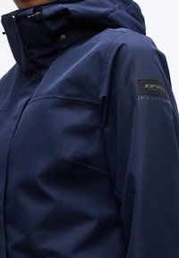 Gros plan d'une personne portant une veste imperméable bleu marine avec un col haut et un patch logo Icepeak sur le haut de la manche.