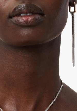 Collana in argento su una pelle scura e liscia, abbinata a un orecchino geometrico con frange metalliche lunghe.
