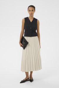 Gilet senza maniche nero con bottoni, abbinato a una gonna crema plissettata. La modella tiene una pochette nera e indossa scarpe nere a punta.