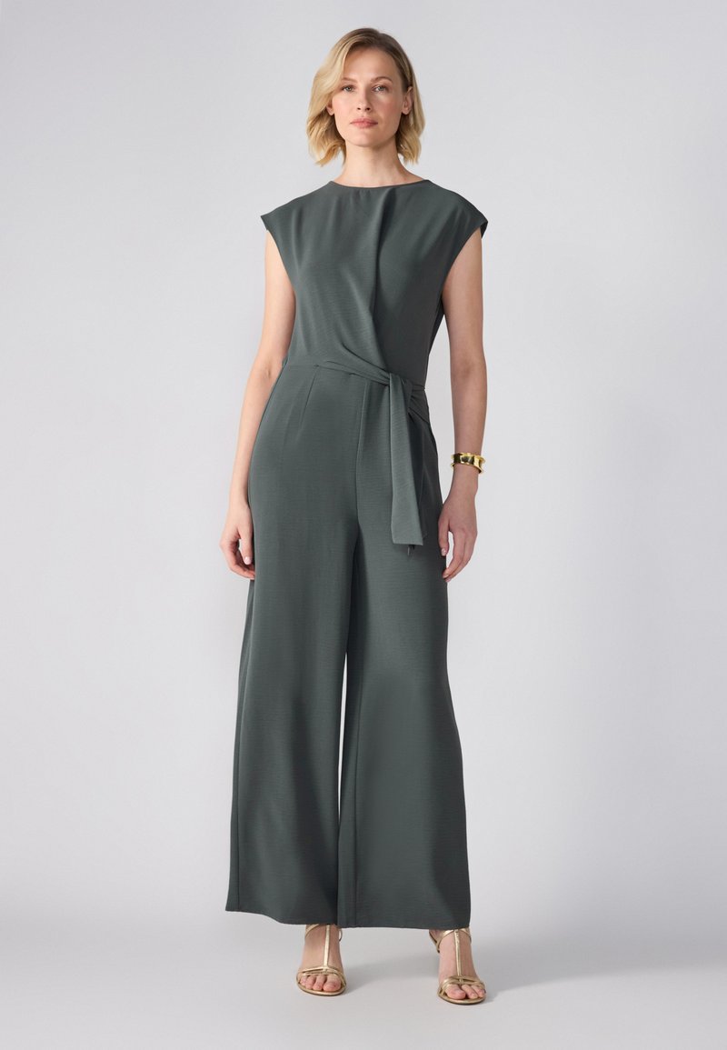 Jumpsuit verde scuro con maniche a cappuccio, design avvolgente con un nodo in vita e gamba larga. Abbinato a sandali dorati con cinturini.