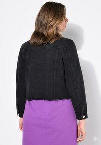 Svart denimjacka med kort längd och fransad nederkant, långärmad och med knäppta manschetter, som bärs över ett lila plagg. Minimalistisk design och textur.