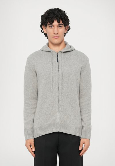 Jeune homme avec des cheveux bouclés et foncés, portant un pull à capuche zippé gris clair et un pantalon noir, se tenant devant un fond blanc uni.