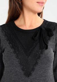 Maglione nero con un'applicazione in pizzo trasparente a V sul colletto, dotato di un nastro nero. Corpo grigio testurizzato.