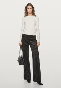 Hvid langærmet top, sorte corduroy bukser med vidde og sort håndtaske. Modellen har spidse hæle på, stående på en neutral baggrund.