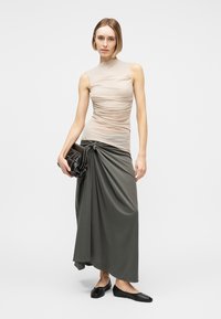 Woman wearing sleeveless beige ruched top and long dark gray draped skirt, holding black chain-strap clutch, with black flats.