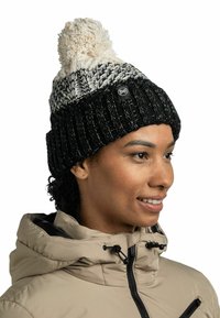 Gorro de punto en blanco y negro con un pompón crema. Presenta un borde acanalado y un parche con logo. Textura suave y diseño cálido.