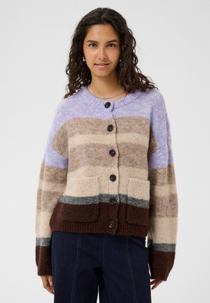 Femme aux cheveux bouclés et foncés portant un cardigan tricoté rayé boutonné violet, beige, gris et marron, associé à un pantalon bleu foncé.
