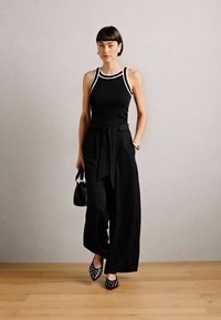 Top de tirantes negro acanalado con ribete blanco, pantalones anchos negros con cinturón de lazo, bolso negro y zapatos de tacón con lunares, todo frente a un fondo neutro.
