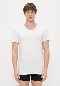 Λευκό κοντομάνικο μπλουζάκι με V λαιμόκοψη, κατασκευασμένο από ελαφρύ ύφασμα, με απλό σχέδιο και άνετη εφαρμογή, σε συνδυασμό με σκούρες βερμούδες.