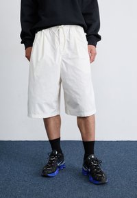 En person står på et blåt tæppe, iført en sort sweatshirt, hvide shorts og sorte sneakers med blå såler. En almindelig væg er i baggrunden.