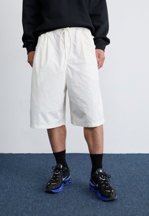 A person stands on a blue carpet, wearing a black sweatshirt, white shorts, and black sneakers with blue soles. A plain wall is in the background.