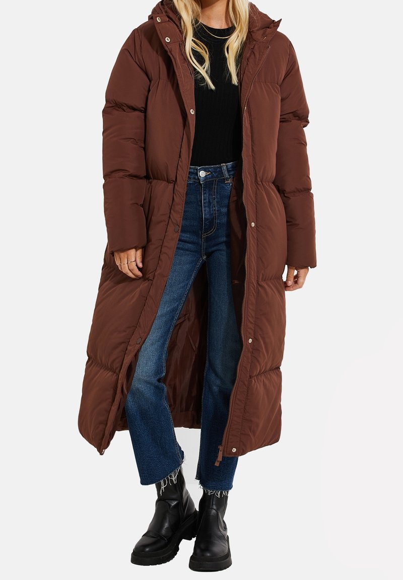Abrigo acolchado marrón con capucha, de largo, con patrón acolchado, cierre a presión y cremallera, combinado con jeans azules y botas negras hasta el tobillo.