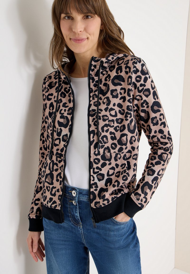 Leopardenmuster-Kapuzenpullover mit Reißverschluss in Beige und Schwarz, mit einer Fronttasche, Kordelzugkapuze und schwarzen Bündchen, kombiniert mit blauen Jeans.