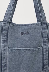 Mørkeblå denim totebag med to robuste stropper, utstyrt med en utvendig lomme og brodert "GAP"-logo på fronten.