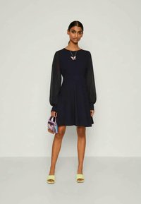 Vestido azul marinho com mangas longas transparentes, saia rodada, combinado com sandálias amarelas e uma pequena mala roxa com um design de borboleta.