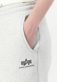 Hand rustend in de zak van lichtgrijze sweatpants met het Alpha Industries-logo gedrukt nabij de zak.