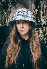 Svart och vit mönstrad bucket hat med breda brätten, bärs över långt, brunt hår. Åtföljd av en svart hoodie, med en texturerad bakgrund.