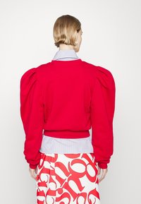 Sudadera roja con mangas abullonadas, puños y ribete acanalados. Usada sobre una camisa azul y blanca a rayas. Fondo blanco liso.