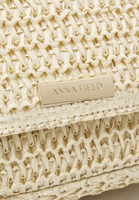 Wiklinowa torebka w kolorze kremowym o teksturowanej powierzchni. Posiada złocistą tabliczkę z logo z grawerowanym tekstem oraz usztywnioną formę.