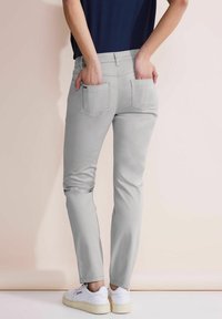 Los pantalones gris ajustados cuentan con bolsillos traseros y una textura suave, combinados con zapatillas blancas sobre un fondo claro.