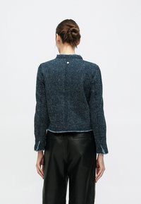 Giacca blu con trama, maniche lunghe, design corto e orlo sfrangiato. Presenta un piccolo dettaglio metallico al collo e spacchi laterali in tessuto.