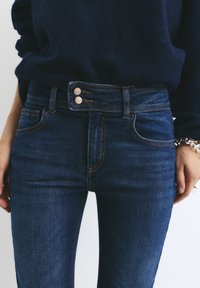 Temno modri kavbojki z visoko pasnico, z dvojnim gumbom, oranžnim šivom in sprednjimi žepi. Gladka tekstura s slim fit oblikovanjem.
