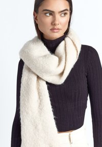 Kobieta ubrana w czarny żebrowany golf i duży, miękki biały szalik luźno zarzucony wokół szyi na jednolitym tle.