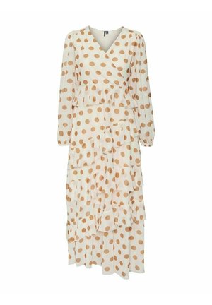 Femme portant une longue robe beige à pois marron, avec un col en cache-cœur, des couches à volants étagées et des manches longues transparentes, debout sur un fond rose.