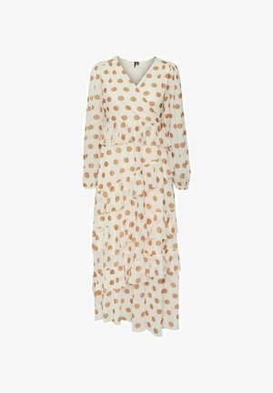 Kobieta w długiej beżowej sukience w brązowe kropki, z dekoltem kopertowym, piętrowymi falbanami i przezroczystymi długimi rękawami, stojąca na różowym tle.