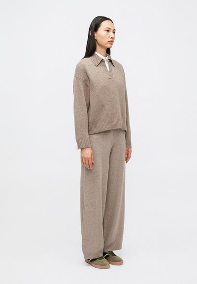 Naine seisab, kandes taupe-tooni kudumit kraega, sobivaid laiade säärtega pükse, valget särk all ja oliivrohelisi spordijalatsid.