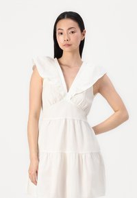 Balta medvilninė suknelė su giliu V formos dekoltė ir raukiniais ant pečių. Sijonas turi pakopinį dizainą, kuris prideda apimties ir tekstūros.