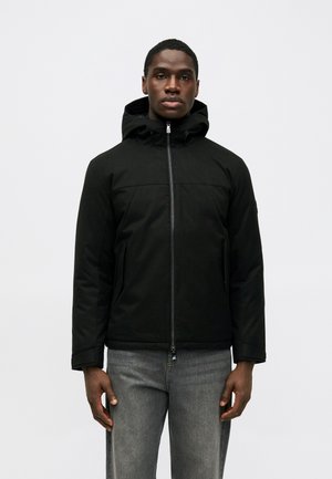 Mustaga kapuutsiga jakk, millel on ees lukk ja kaks küljetaskut, valmistatud tekstureeritud kangast, seljas hallide teksade peal.