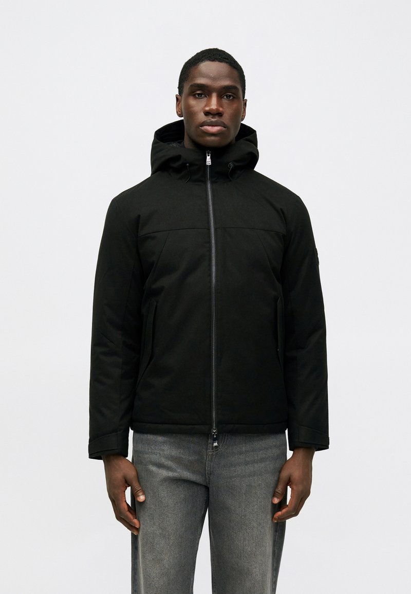 Casaco preto com capuz, apresentando um fecho à frente e dois bolsos laterais, feito de tecido texturizado, usado sobre jeans cinza.