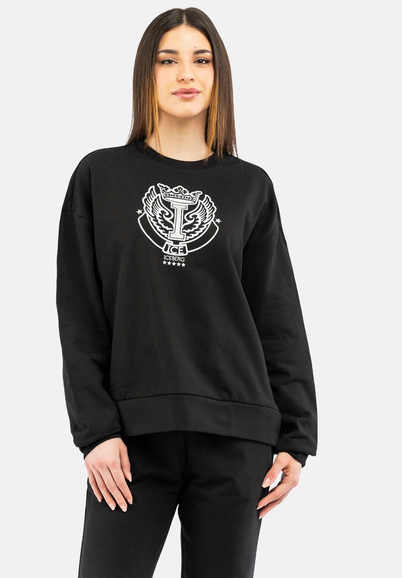 Svart sweatshirt med rund halsringning, oversized passform och vit grafisk emblem på framsidan. Långa ärmar och ribbade cuffar vid nederkanten.