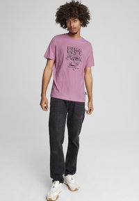 Rosa Baumwoll-T-Shirt mit einem schwarzen Grafikdruck, kombiniert mit schwarzen Jeans und weißen Sportschuhen. Das Model steht vor einem neutralen Hintergrund.