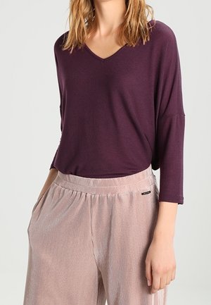 Kvinde iført en løs, langærmet lilla V-hals top, som er stoppet ned i lys pink, vidtløftede bukser med høj talje og lommer.