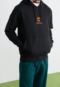 Sudadera negra de algodón, con un bolsillo frontal y un logo naranja de Timberland. Usada con pantalones color teal; tela texturizada visible.