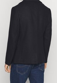 Sort, skræddersyet blazer lavet af en uldblanding, med en klippede krave, slank silhuet og sidelommer, parret med denimjeans.