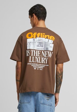 Brown oversized t-shirt featuring white and orange text on the back, graphic of vintage technology, and soft cotton fabric.