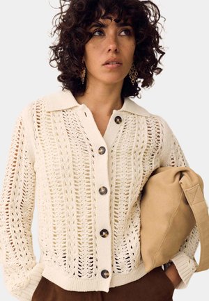 Vrouw met krullend donker haar, die gouden oorbellen draagt en een crèmekleurige gebreide cardigan, houdt een beige tas vast tegen een effen achtergrond.