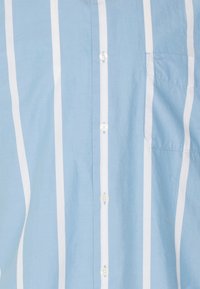 Camisa azul claro con rayas blancas verticales; presenta un bolsillo en el pecho y botones de plástico. Hecha de una tela suave y texturizada.