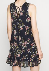 Robe sans manches florale bleu marine foncé avec des motifs blancs, roses et verts. Présente un ourlet à volants et une fermeture par lien à l'arrière.