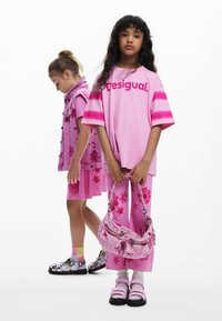 Dve deti v ružových odevoch. Dievča vľavo má na sebe ružové kvetinové šaty; dievča vpravo má ružovú košeľu a voľné nohavice, drží kabelku.