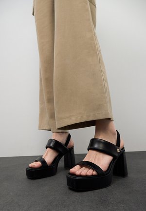 Black platform sandals with wide straps, open toe, and a chunky block heel. Designed with a smooth leather texture and gold hardware accent.